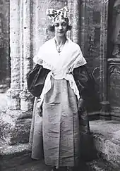 Photographie noir et blanc d'une jeune femme portant une robe à manches bouffantes, un châle blanc, et une coiffe avec un nœud