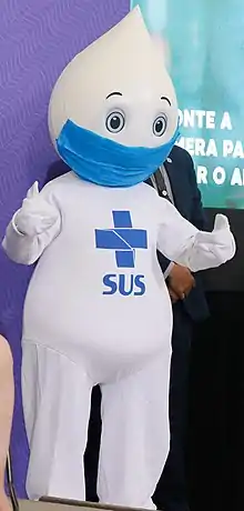 Personnage habillé de blanc, au visage en forme de goutte, portant un masque chirurgical bleu. Sur sa poitrine est inscrite une croix bleue et les lettres SUS représentant le système de santé brésilien