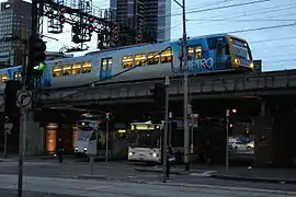 Un tramway à côté d'un bus Melbourne Bus Link, avec un métro Trains X'Trapolis 100 passant au-dessus