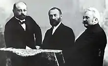 Noir et blanc, les trois sont habillés en costume noir avec chemise blanche et nœuds papillons noirs. Les deux de gauche ont une barbe et sont bruns, celui de droite à une moustache et des cheveux blancs.