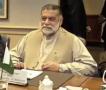 portrait d'un homme barbu assis devant un table, et devant lui, un petit drapeau pakistanais de table