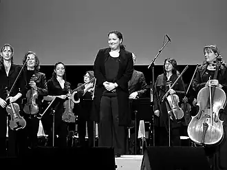 Photographie en noir et plan d’une femme debout, se tenant devant quelques musiciens.
