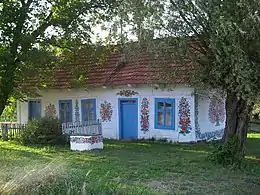 Photographie couleur d'une petite maison blanche aux volets bleus, cernée d'arbres et sur les murs de laquelle sont peints des motifs floraux.