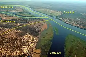 Vue aérienne du Zambèze, à la jonction entre : • la Namibie en haut à gauche, entre le Zambèze et son affluent le Kwando constitue la pointe de la bande de Caprivi, • la Zambie, en haut, au-delà du Zambèze, on aperçoit sur la rive du fleuve le faubourg de Kazungula, • le Zimbabwe en bas à droite avec l'île et une partie de la rive sud du Zambèze, • le Botswana en bas à gauche, sur la partie droite de la rive. On distingue au fond le tracé de la route reliant Kasane au ferry menant à Kazungula, remplacé depuis 2017 par un pont.