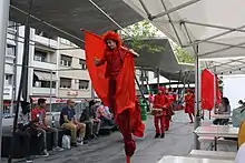 Parade rouge comprenant des échassiers, des danseurs et des musiciens percussionnistes.