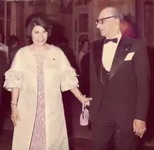 Une femme en robe rose et manteau blanc tient la main d'un homme en costume-cravate.