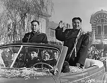 Photographie montrant deux hommes paradant à l'arrière d'une voiture décapotable, Kim Il-sung est à droite et salue la foule.