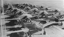 Environ trente appareils, dont certains sont bâchés, se trouvent sur le pont d'un porte-avions. Des personnels au sol sont assis à l'ombre des ailes.