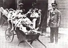 Une charrette est remplie de cadavres squelettiques d’enfants. Trois hommes l’entourent, dont au moins un porte une casquette de policier ; un autre porte un cadavre dans ses bras.