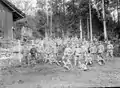 Photographie noir/blanc d’hommes, en uniforme militaire pour la plupart, posant en groupe près d’une grange en bois et devant une forêt