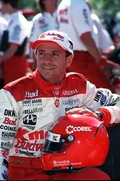 Photo d'un homme souriant, assis, vêtu d'une combinaison de course blanche et rouge, son casque de course rouge posé sur sa cuisse gauche