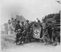 Groupe de fantassins alignés devant un char allemand naufragé et affichant une croix gammée capturée.