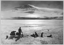 Photo en noir et blanc de deux hommes et cinq chiens attachés à un traîneau à l'arrêt sur la banquise devant un volcan.