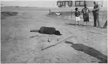 Photo en noir et blanc avec un corps étendu de dos sur un sol aride, trois personnes discutant en second plan sur la droite.