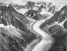 Photo aérienne en noir et blanc d'un glacier en S, présentant des stries en V, au pied de sommets abrupts.