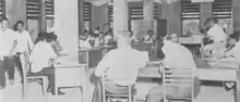 Photo en noir et blanc d'hommes assis sur des chaises, derrière des bureaux formant un cercle.
