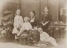 Marie comtesse de Flandre, assise sur un fauteuil capitonné et vêtue élégamment pose avec ses quatre enfants : Henriette, Albert et Joséphine vêtus de tenues claires et Baudouin en costume avec une cravate