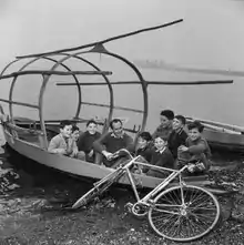 Photographie en noir et blanc de Koblet assis dans une barque au milieu d'enfant, sur les rives d'un lac, son vélo posé devant.