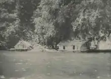 Photo en noir et blanc montrant des habitations sur pilotis au pied d'une falaise.