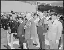 Un homme asiatique et un groupe d'hommes blancs, se tenant debout sur un terrain d'aviation militaire, dans une position de recueillement.