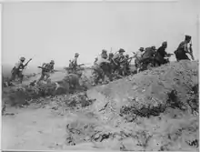 Photographie d'un groupe de soldats en uniformes légers et portant des chapeaux de feutre courant vers le sommet d'un talus la baïonnette en avant