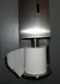 Distributeur vertical de papier toilette, avec déroulement du rouleau dans le sens anti-horaire.